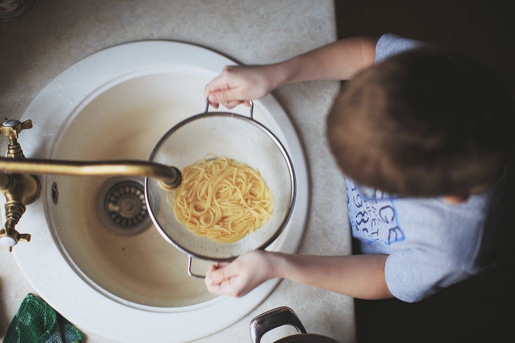 A child holding a strainer with pasta over the sink.