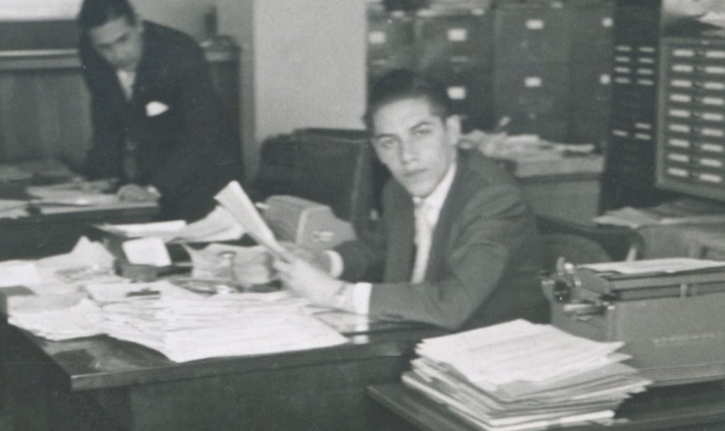 My father at his desk, surrounded by papers and an antique typewriter. Colleague and file cabinets in the background.