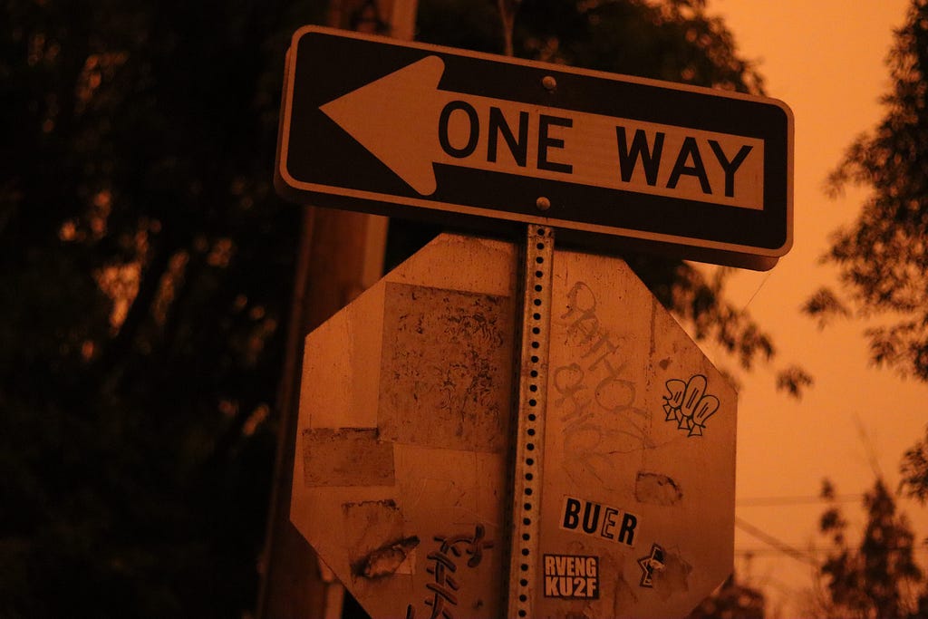 The back of a STOP sign and the front of a ONE WAY pointed left are tinted orange due to smoke from wildfires.