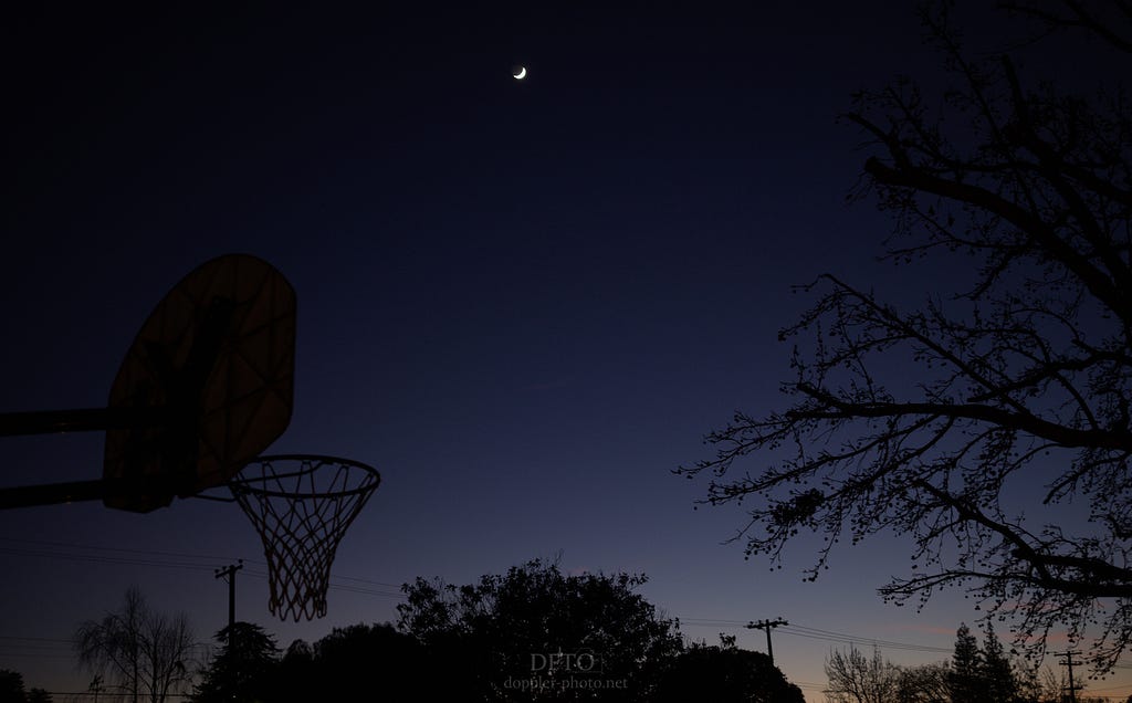 A crescent moon floats high above the last embers of a failing dusk. Silhouettes of trees, power lines, and a basketball hoop anchor the scene to the earth.