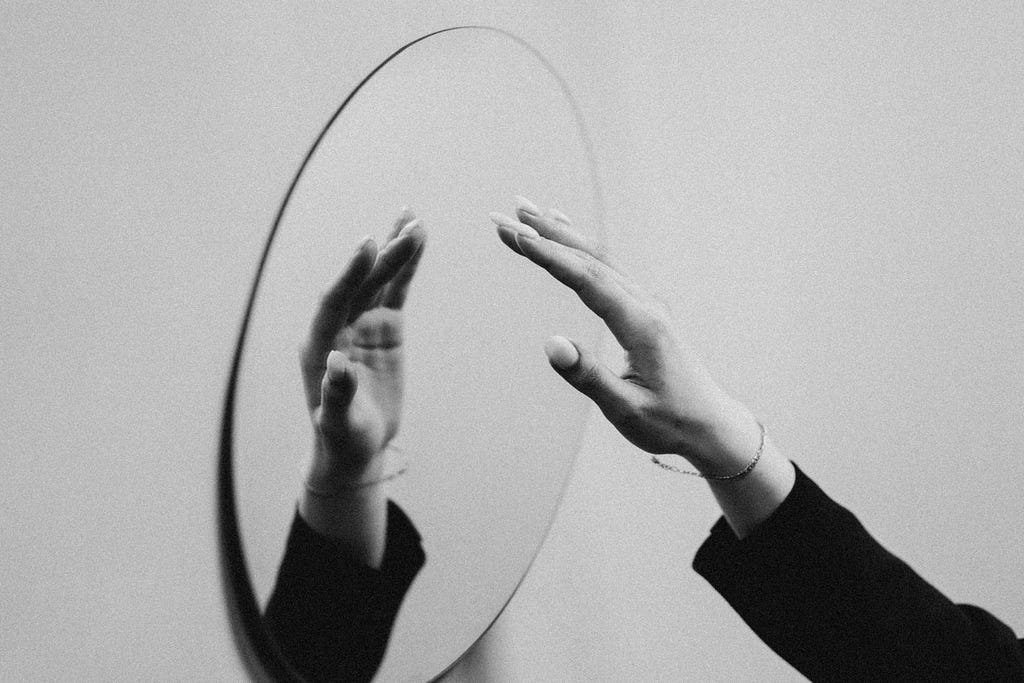 A black and white photo of a mirror on a white wall viewed from a slanted angle. A hand is extended towards the mirror, and its reflection is visible. The hand has white painted fingernails, a thin bracelet, and we can see the top of a black long-sleeve.