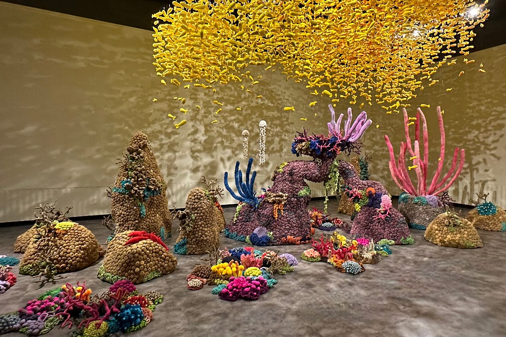 In a museum, a huge school of bright yellow fish hang from the ceiling. Underneath is a sprawling coral reef of all colors, shapes, and sizes. All of it is made up of crochet.
