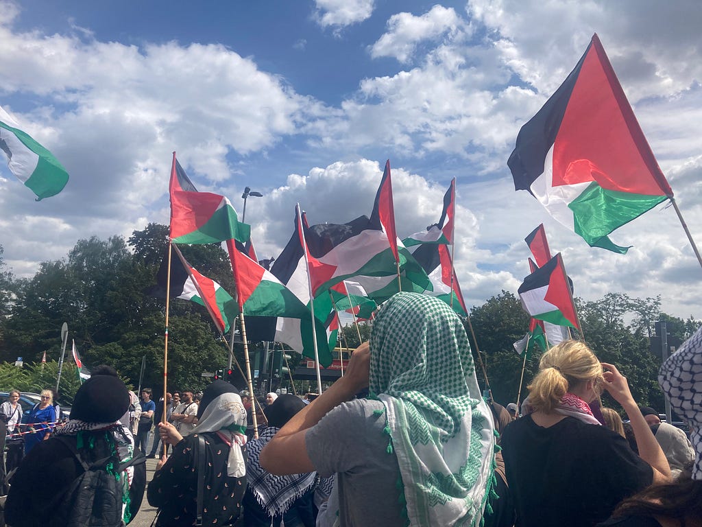 A protestor wearing a green keffiyeh stands with his hands cupped around his mouth amidst Palestinian flags