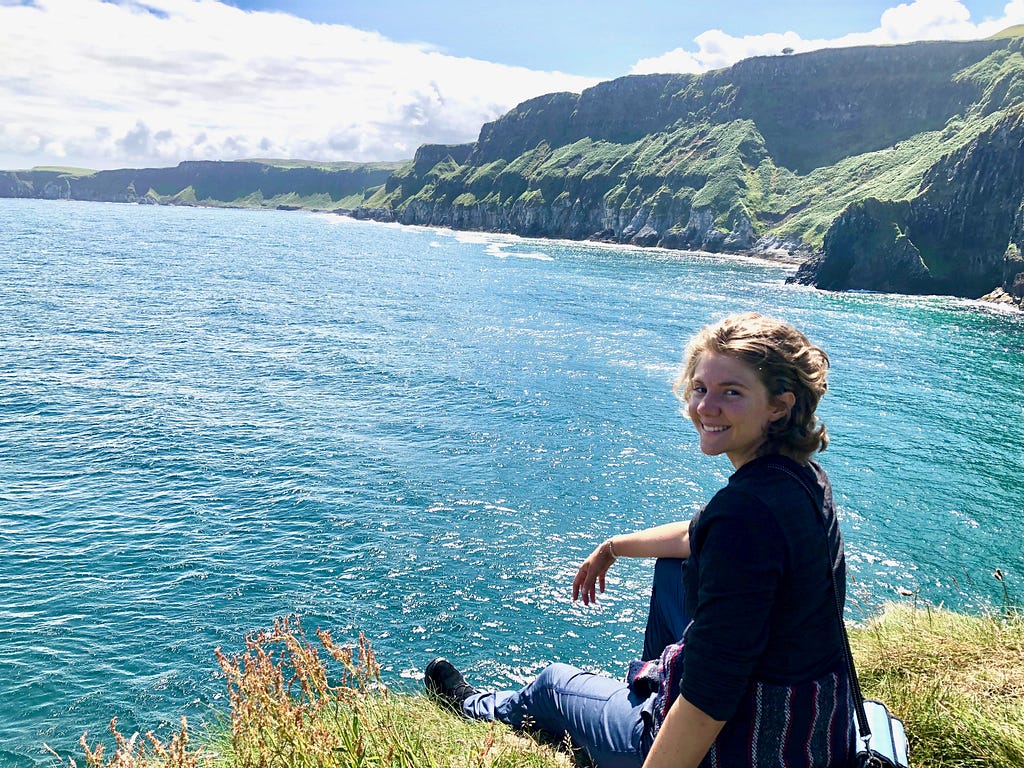 A woman — Hannah Brown — smiles at the camera while sitting on a cliff overlooking blue water and green cliffs.