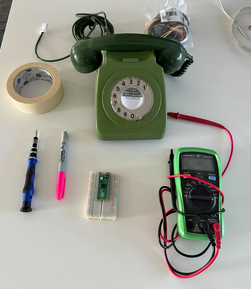 Green rotary telephone, masking tape, flat-head screwdriver, sharpie, rasperry pi pico, and multimeter arranged neatly on a desk