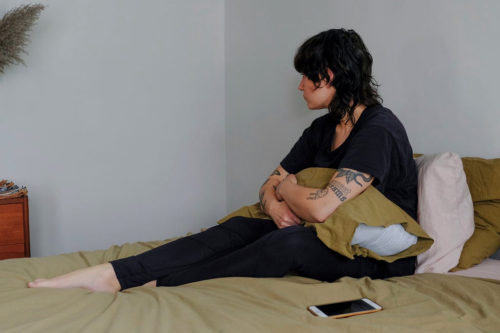 Person wearing all black sits in bed with a pillow on their lap and looks away from the camera.