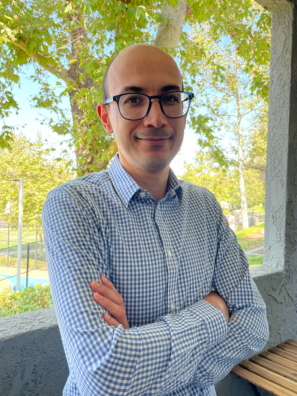 Whittier College’s own lecturer, José Contreras stands, arms crossed in front of a view of a tree. He wears a a blue and white checkered collared shirt, glasses and wears a half-smile.