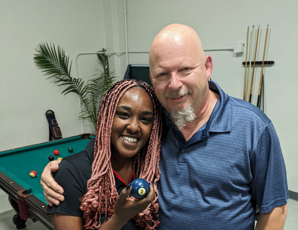 Partners Amani and Doug pose for a picture in front of a pool table while holding the 2 ball