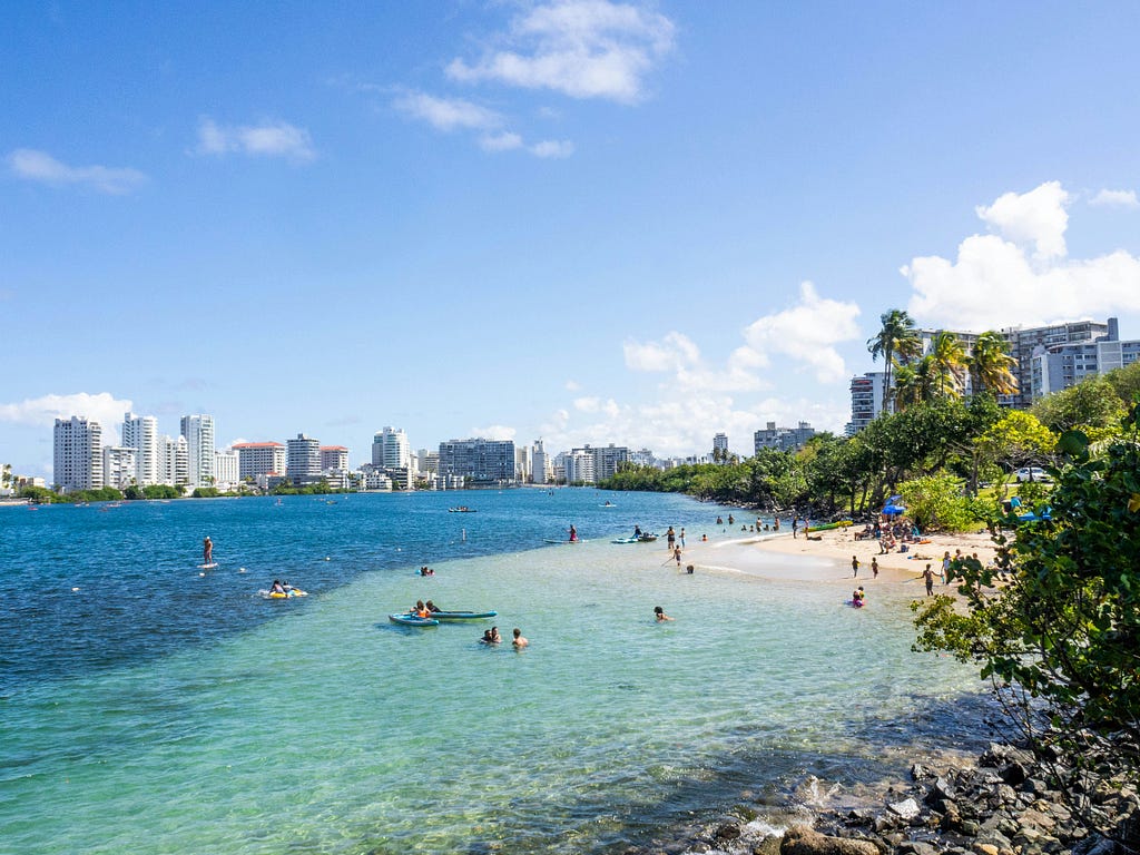 View of the beach in Condado light turquoise water and darker blue. It is surrounded by condo buildings and a blue sky.
