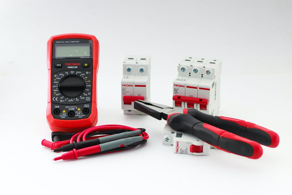 A red digital multimeter, red cords and prongs, red pliers, and other tools on a while background