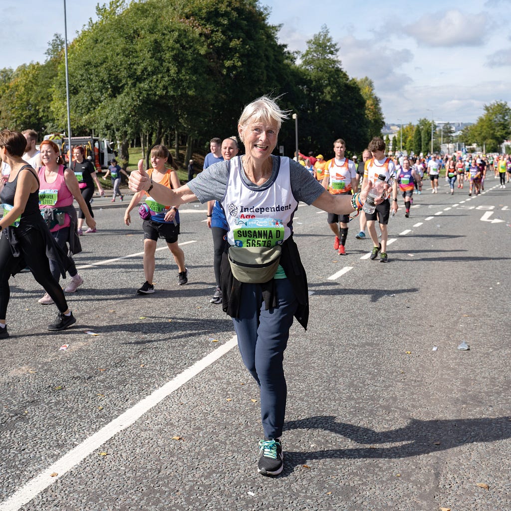 An older woman smiles at the camera as she runs. She holds her hands in a thumbs up and is wearing an Independent Age running vest