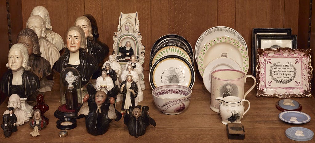 Various different John Wesley ceramics on display on a wooden shelf, including figurines of the Methodist leader, plates, jugs, bowls and effigies.