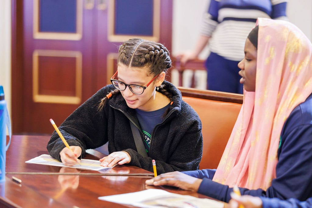 Two students write on paper while sitting at a table.