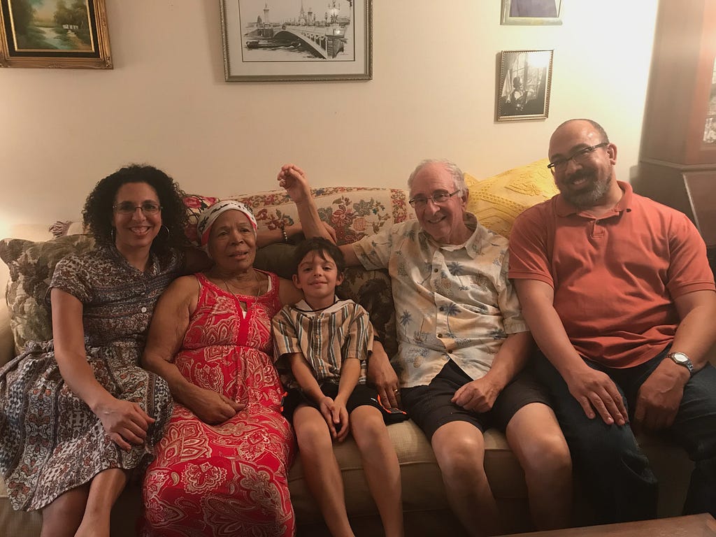 Five people seated on a sofa, from left: a light-skinned 50 year old biracial woman wearing a blue patterned dress with her left arm behind a 78 year old Black woman wearing a red dress; whose left arm is around a 9 year old white-appearing boy wearing a vertical-striped shirt; next to a 78 year old white man in a white patterned shirt with his right arm at a right angle behind the boy’s head; next to a tall, biracial man wearing a burnt orange shirt. They all smile at the camera.