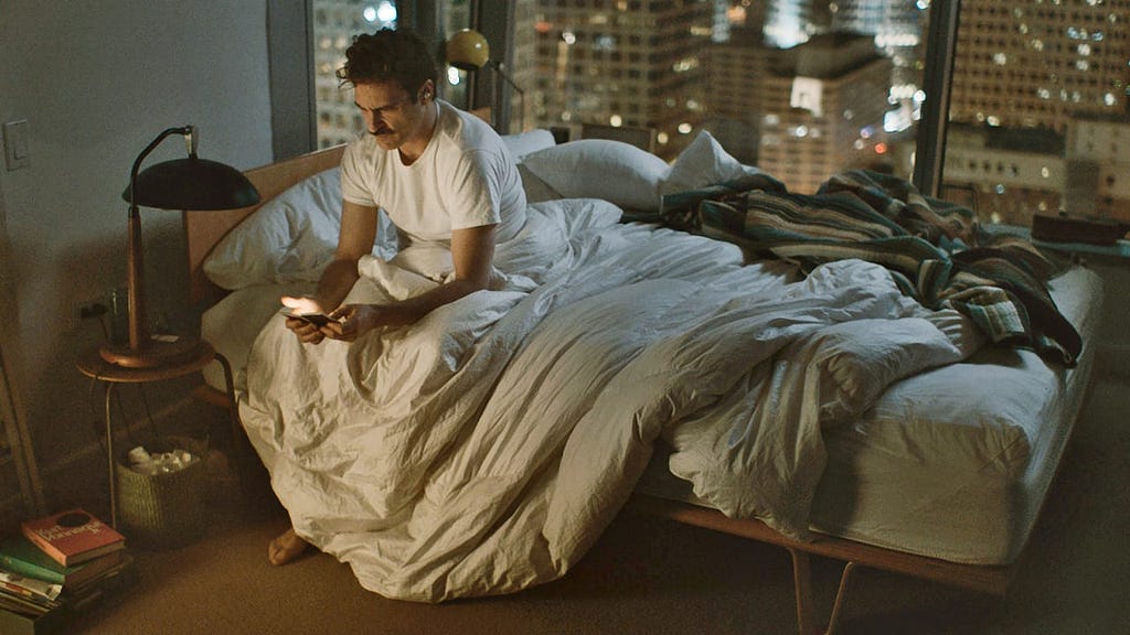 Snapshot of Spike Jonze’s movie Her, where Joaquin Phoenix sits alone on his bed, with a phone in his hand and a sad face