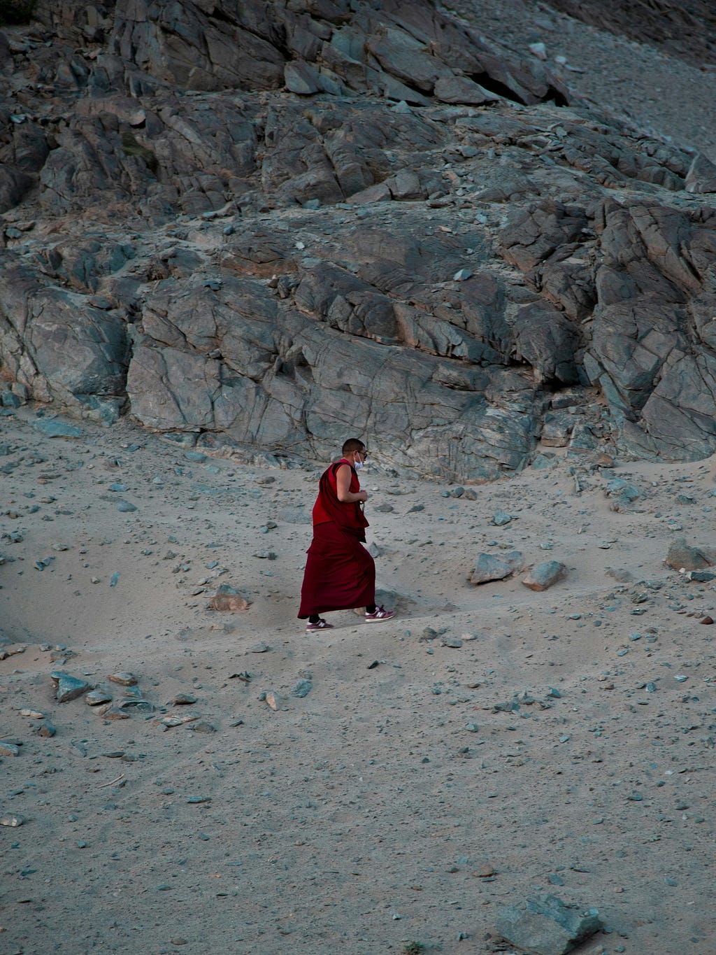 Tibetan Buddhist monk wearing sneakers and a long maroon robe walking on a grey dusty path — rocks are scattered around the path and there is a big rock hill in the background