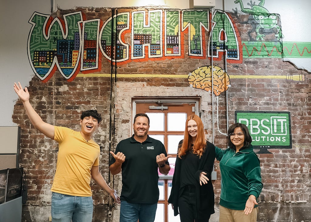 Three young people and one IT professional pose joyfully in front of a mural that says “Wichita.”