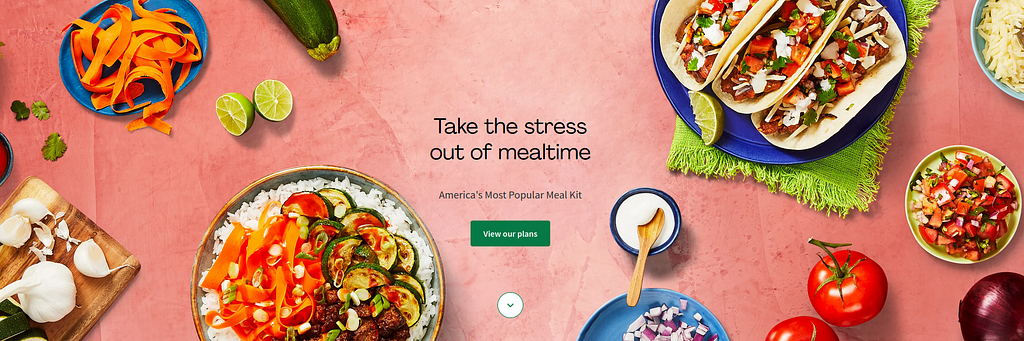 A salmon pink background (table top) showing meals such as colourful tacos with cheese and peppers, a bowl of salsa, and pan fried zucchinis over rice. Ingredients tastefully scattered around, such as a zucchini, a tiny bowl of mayo, tomatoes, garlic, onions, limes.