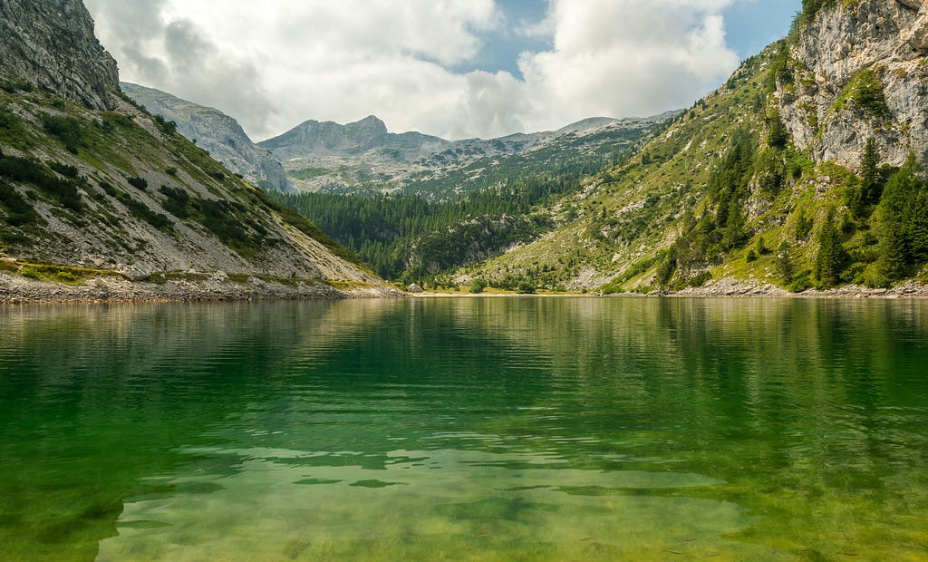 A lake surrounded by mountains, hills, and forests. The lake water reflects the green of its surroundings.