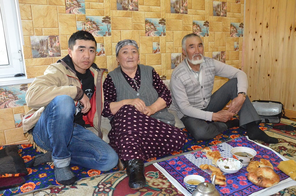Son, mother, and father sit on a carpeted floor with a meal and tea laid out in front of them.