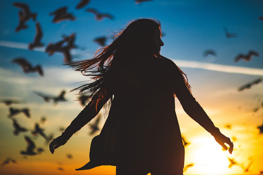 A swirling woman is joined by a flock of birds against a blue-orange backdrop of a sunrise.