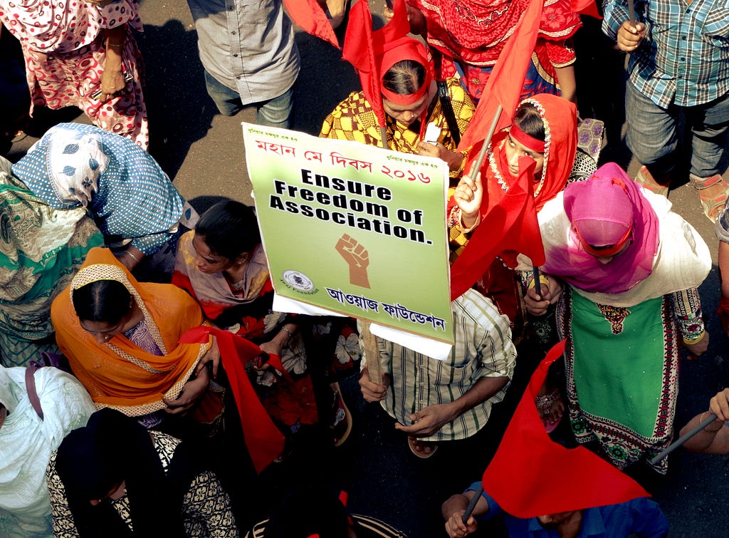 A group of women in colorful dresses and head coverings as well as a few men dressed in typical street clothes assemble at a protest. One person holds a placard with a raised fist in the center, and with Bengali text and English text that says “Ensure Freedom of Association.”