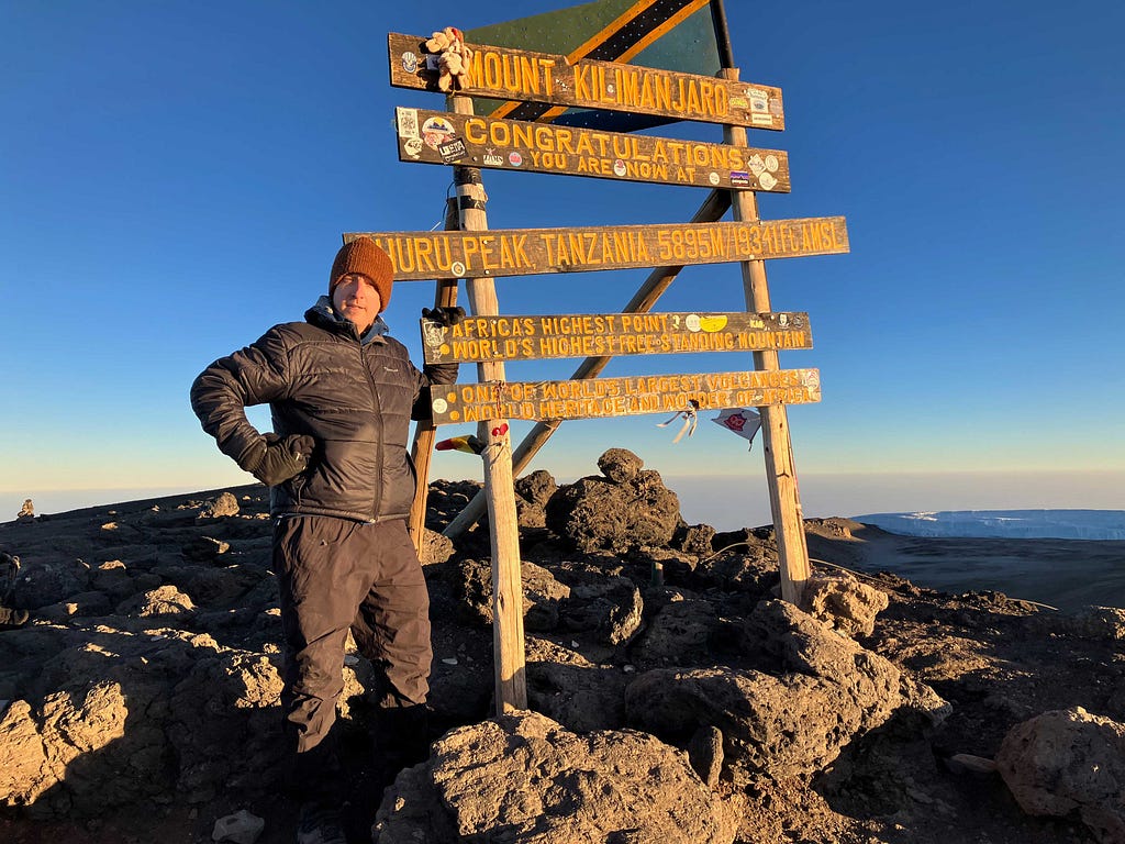 The author pictured standing amid a collection of boulders beside the summit sign at Uhuru Peak on Mount Kilimanjaro under deep blue sky.