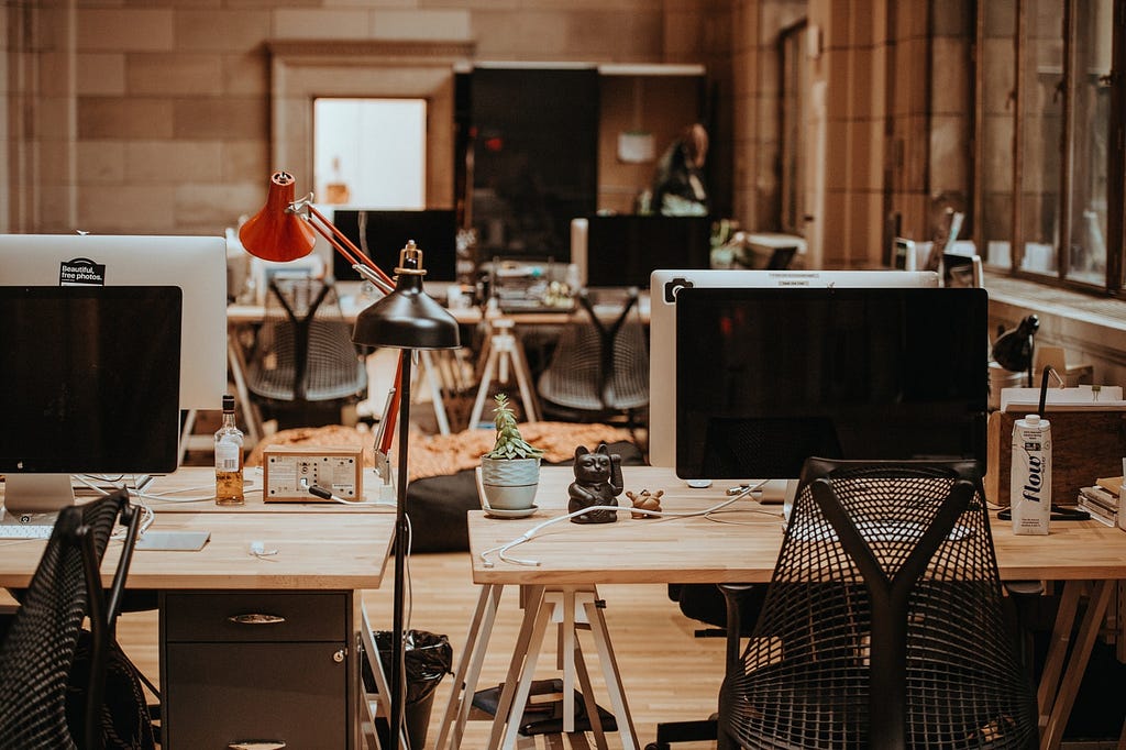 An office space, devoid of people, with several wooden desks sporting Apple monitors and personal effects