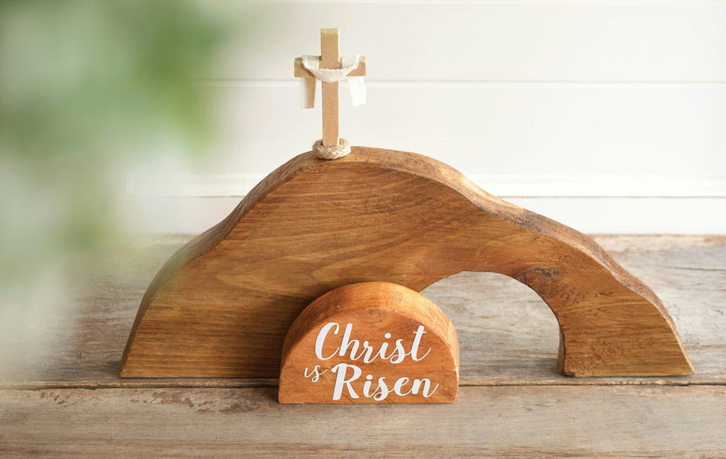 A wooden sculpture art with a mini cross on top, and a white-painted script that says “Christ is Risen”.