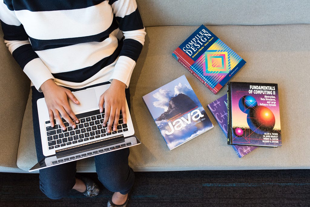 Mulher segurando um computador no colo e livros de programação espalhados ao seu lado.