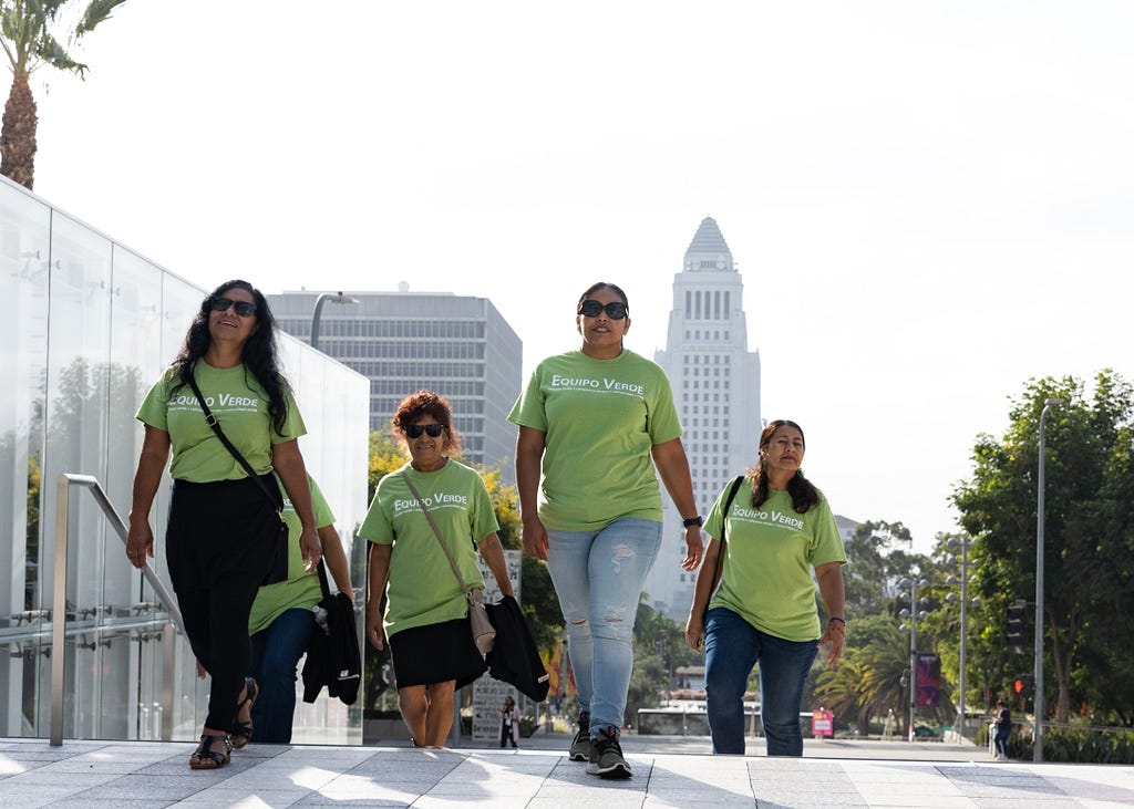 Four women in matching green t-shirts reading “Equipo Verde” and sunglasses walk up the steps in front of LA City Hall.