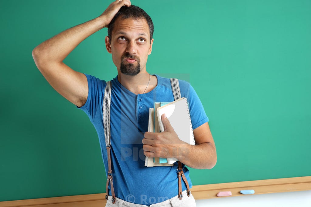 An adult man standing in front of a chalkboard wearing suspenders holds a pile of papers and books in one hand and scratches his head with his other hand.