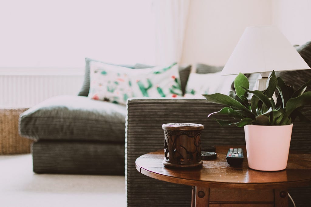 A picture of a livingroom. Wooden side table with a plant in a pot, a remote, a lamp, and a candle on it. Next to the table is an out-of-focus couch with patterned throw pillows.