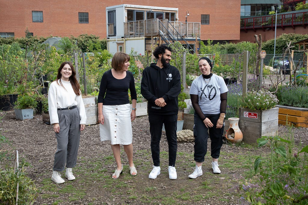 Four people standing in a community garden. The first person on the left person has long straight brown hair and is wearing a white sweater and grey dress pants. The second person has short straight brown hair and is dressed in a black long-sleeve and a white knee-length skirt. The third person has curly black hair and is dressed in a black long-sleeve and black pants. The fourth person is wearing a blue head scarf, a grey shirt over a brown long sleeve and black pants. All of them are smiling.