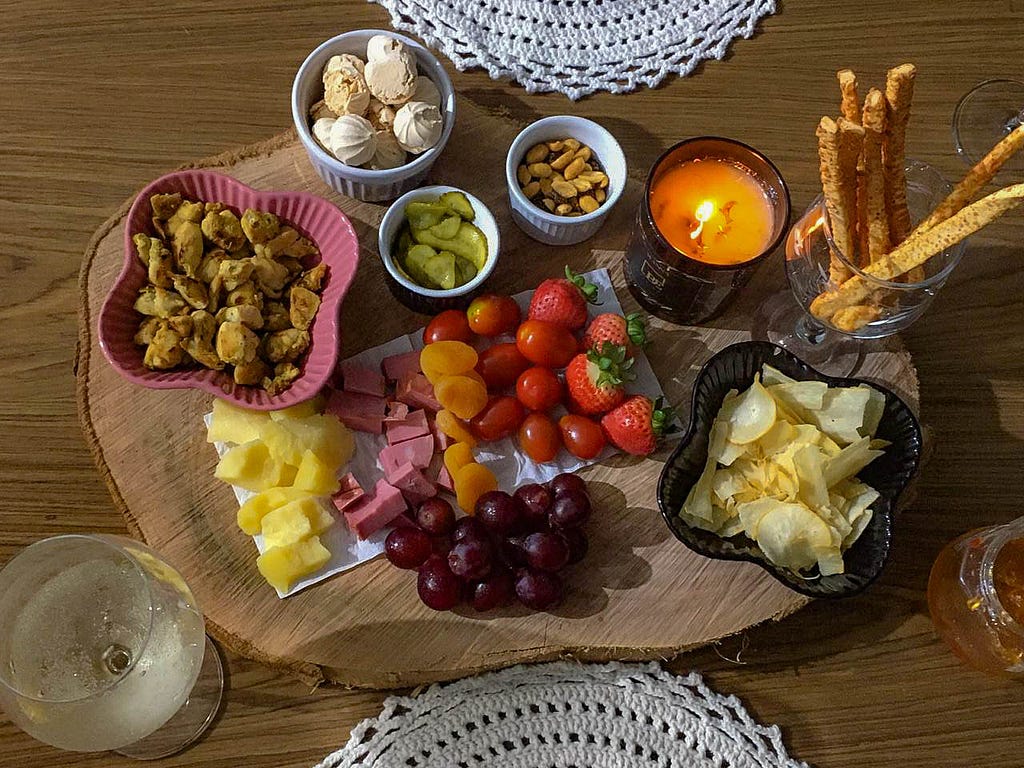 Tábua de frios e frutas: morangos, uvas, queijos, salame, chips de batata, amendoim, tomate cereja. Há uma vela acesa.