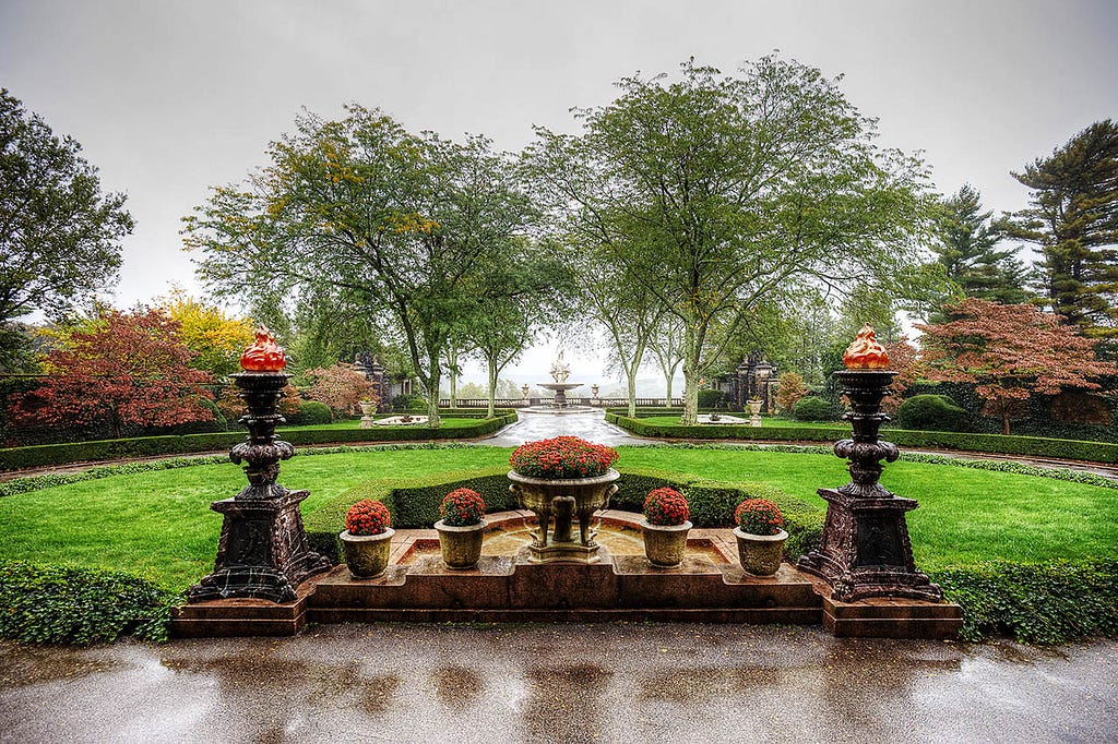 A lush green garden with a tiered fountain in the center. The fountain’s water cascades down into a pool below. In the background, tall trees can be seen.