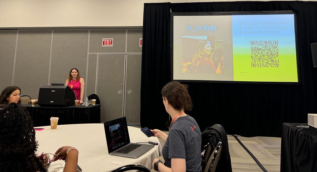 Kelly Vaughn hosting a workshop in a classroom setting with a large projector and attendees sitting in groups around tables with laptops.