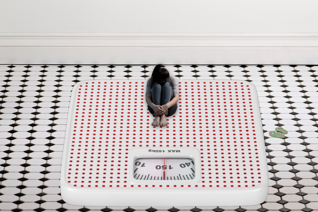 A small-sized girl sitting on a giant weighing scale and feeling depressed.