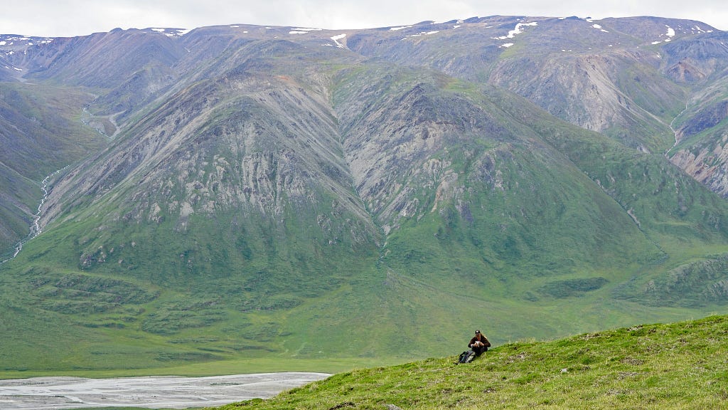 A distant photo of a person sitting on a green hillside overlooking mountains and a river.