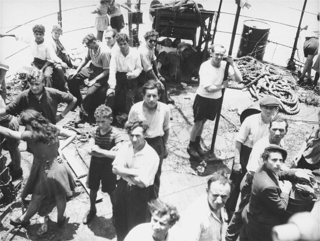 Approximately 20 passengers, including men, women, and children, stand on the deck of the Exodus 1947 ship. The ground is covered with debris and rope.