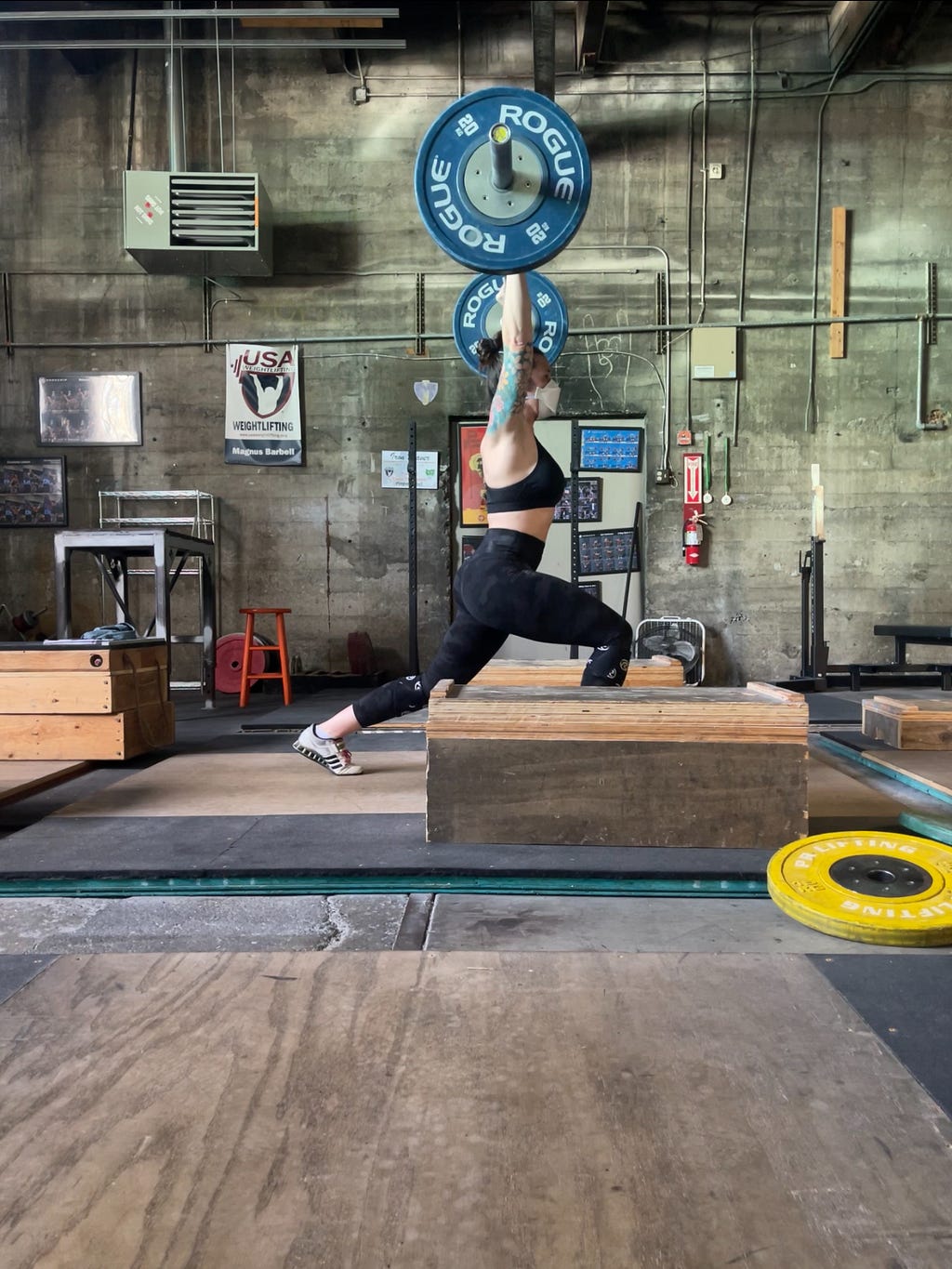 The article author, Kelsey, a white autistic barbell athlete, split jerking 55 kilograms in an empty gym. She’s wearing old-school Adipower lifting shoes, black leggings, a black sports bra, and a white Aura 3M respirator. She’s surrounded by weightlifting platforms, bumper plates, and jerk blocks.