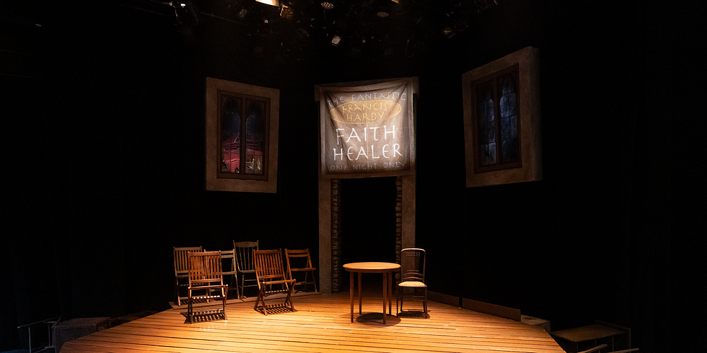 A theatrical set with wooden chairs, wooden floor, and a banner that reads “The Fantastic Francis Hardy, Faith Healer, One Night Only”