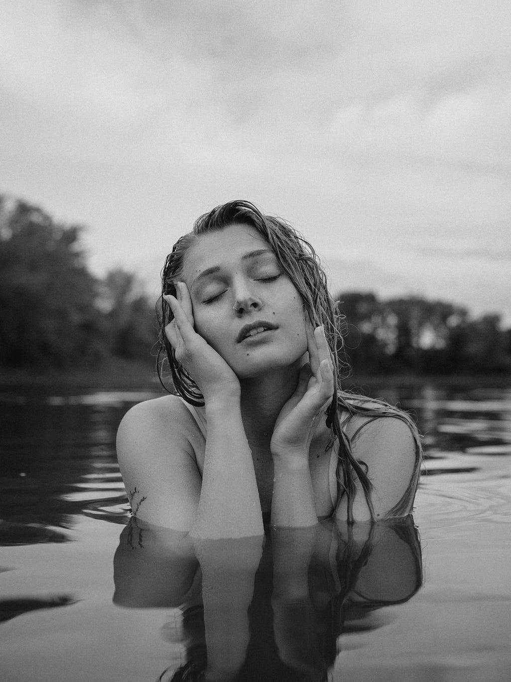 Woman standing in water up to her chest, touching her face with her eyes closed as if daydreaming.