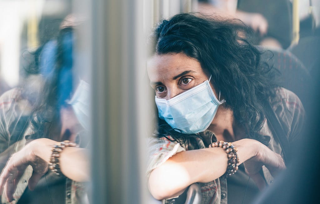 A woman in a surgical mask looks pensively out the window of a bus.