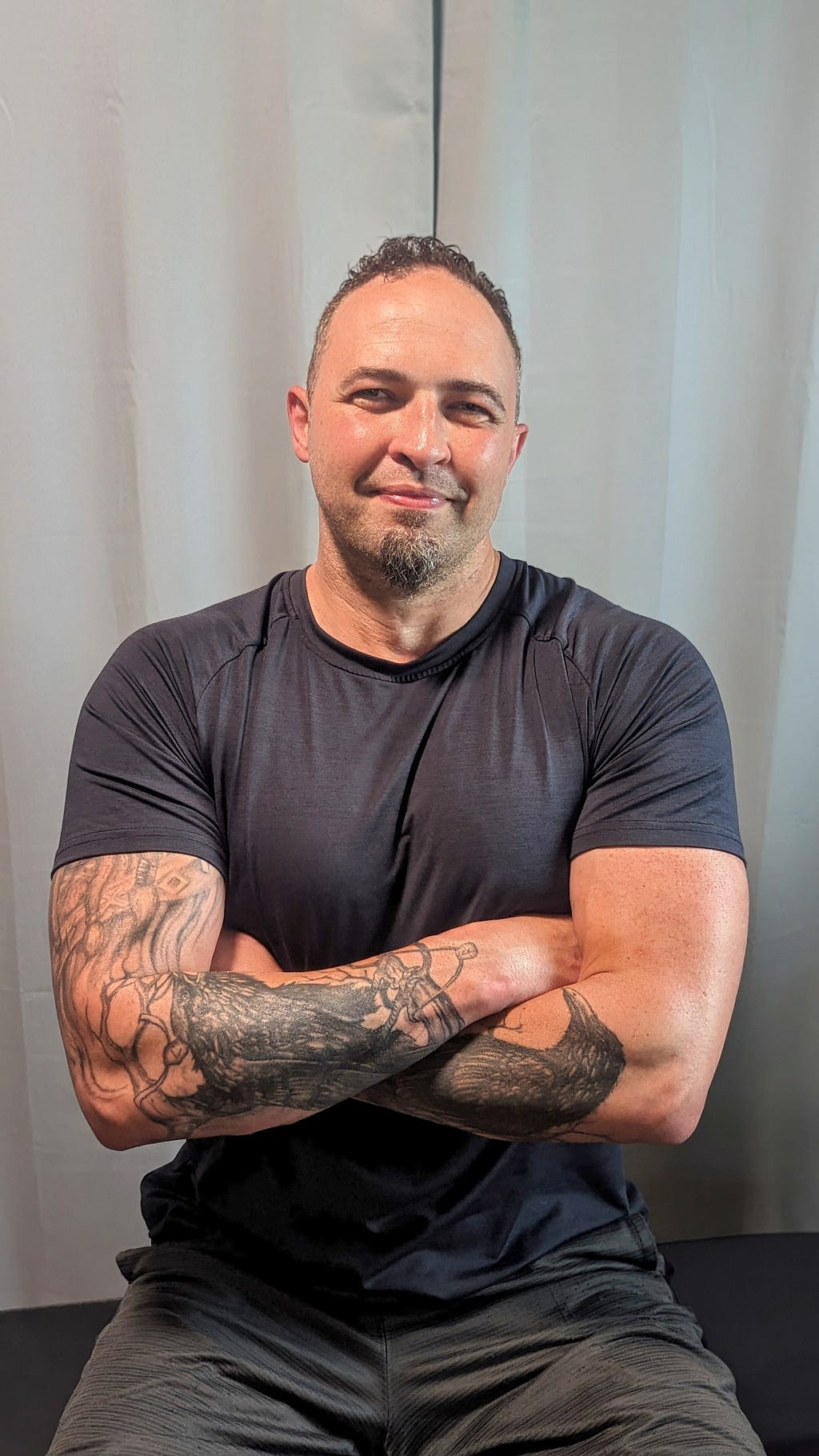 Metahuman Dave is sitting on a workout bench with his arms crossed. Wearing a black T-Shirt. His arm tattoos are visible. Two ravens on his forearms. He is smiling at the camera. This photo was used in his profile picture.