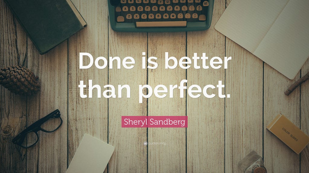 An overhead shot of a wooden desk with part of a typewriter showing, as well as other items which suggest it is a writer’s desk. In the middle is bold white text “Done is better than perfect”, and credits Sheryl Sandberg in smaller pink text underneath the quote.