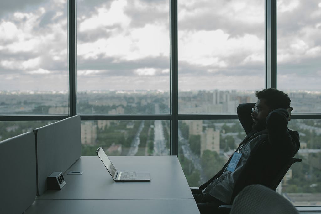 Man sitting and thinking at a desk in a corporate office with large windows and city scape in the background.
