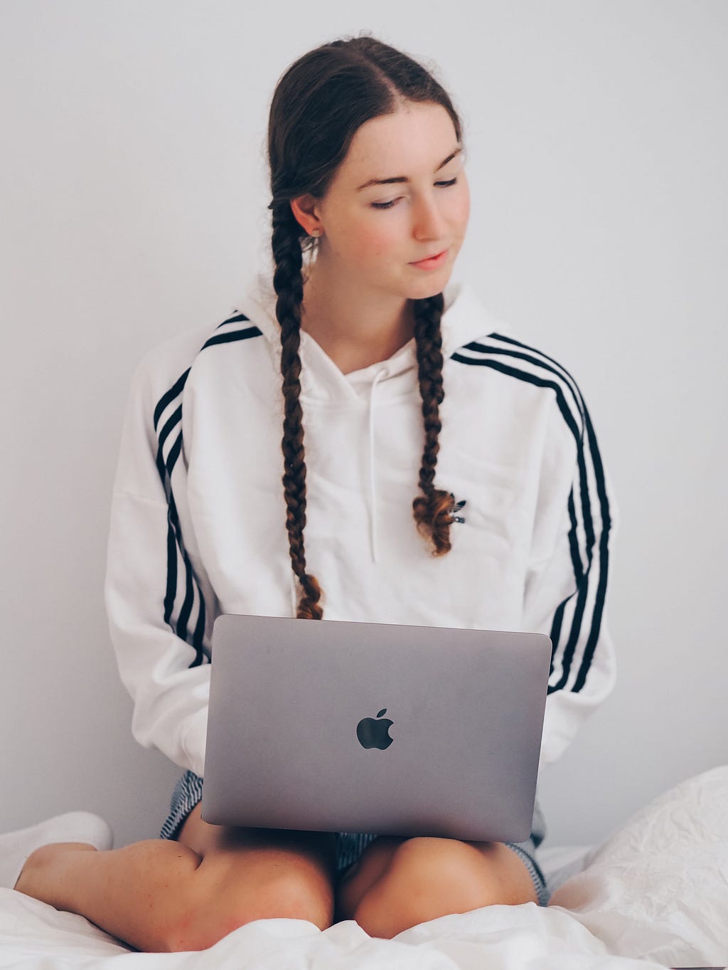 A young white woman with brunette hair styled in two plats either side, wearing a short skirt, small white socks, and a white hoodie with three black stripes running down the top of both sleeves, sitting against a white wall on a white blanket seemingly on top of a bed, with her knees bent and her feet to one side and a Macbook resting on her lap.