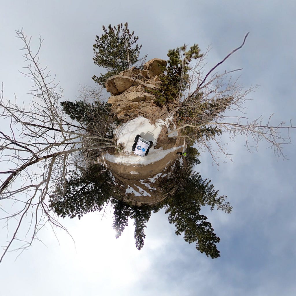 360-degree camera view overhead of the rover in front of the outcrop. The rover is a square robot in the center of the image with four wheels. In front of the rover is an exposed rock outcrop. Behind the rover is a flat wide dirt road. The rest of the landscape is thinly-forested trees with a light coating of snow.