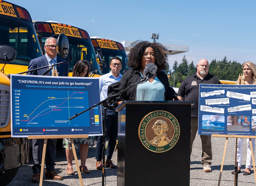 Photo of Yolanda speaking at the podium with the governor and others standing behind her. The electric school buses are visible behind her as well as a poster showing the rising profits of oil companies.